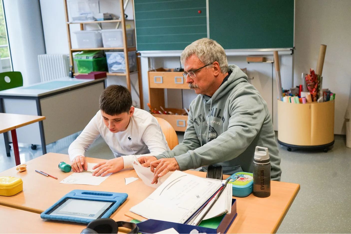 Unterstützungssituation im Klassenraum: Eine Schulbegleitung und ein Kind sitzen an einem Tisch und arbeiten gemeinsam an Arbeitsblättern und Papieraufgaben.